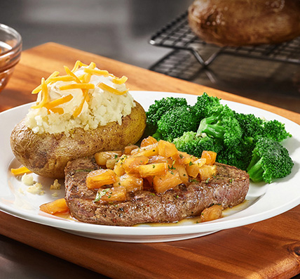 Cheddar's delicious center-cut top sirloin grilled with pineapple and a sweet Hawaiian glaze, served with broccoli and a loaded baked potato on a plate.